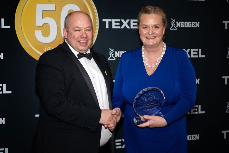 A man and woman shaking hands while posing for a photo. The woman is holding an award.